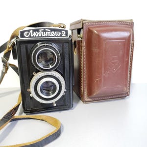 May include: A black twin-lens reflex camera with the text "Любитель 2" on top, featuring two circular lenses. A brown leather case and a leather strap are also visible. The camera is on a light gray surface.