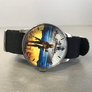 May include: A wristwatch with a black nylon band and a silver-colored case. The watch face features a colorful illustration of a person in a military uniform. The background has yellow, blue, and white hues. The watch hands are gold-colored.
