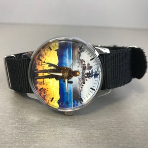 May include: A round wristwatch with a black nylon strap. The watch face features a split design with a soldier silhouette against a yellow and blue background on the left and a cityscape on the right. The watch hands are silver.