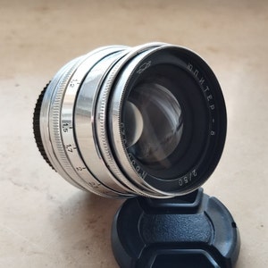 Puede incluir: Un objetivo de cámara plateado con detalles en negro y una tapa de objetivo negra. El objetivo tiene una serie de números y marcas, incluyendo "2/50" y "Jupiter-8". El objetivo está en ángulo, mostrando su cristal reflectante.
