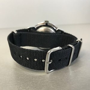 May include: A wristwatch with a black nylon strap and a silver-colored buckle. The watch face is visible, with a silver-colored case. The strap is secured with silver-colored metal loops.