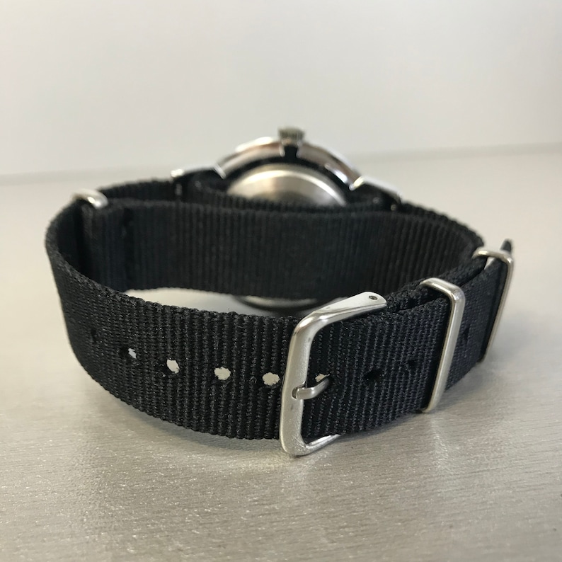 May include: A wristwatch with a black nylon strap and a silver-colored buckle. The watch face is visible, with a silver-colored case. The strap is secured with silver-colored metal loops.