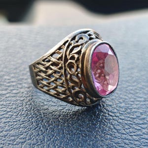 Puede incluir: Un anillo plateado con una gema rosa ovalada. El anillo presenta un intrincado diseño calado en la banda, con un patrón tejido y adornos decorativos. La gema está engastada en un engaste de bisel.