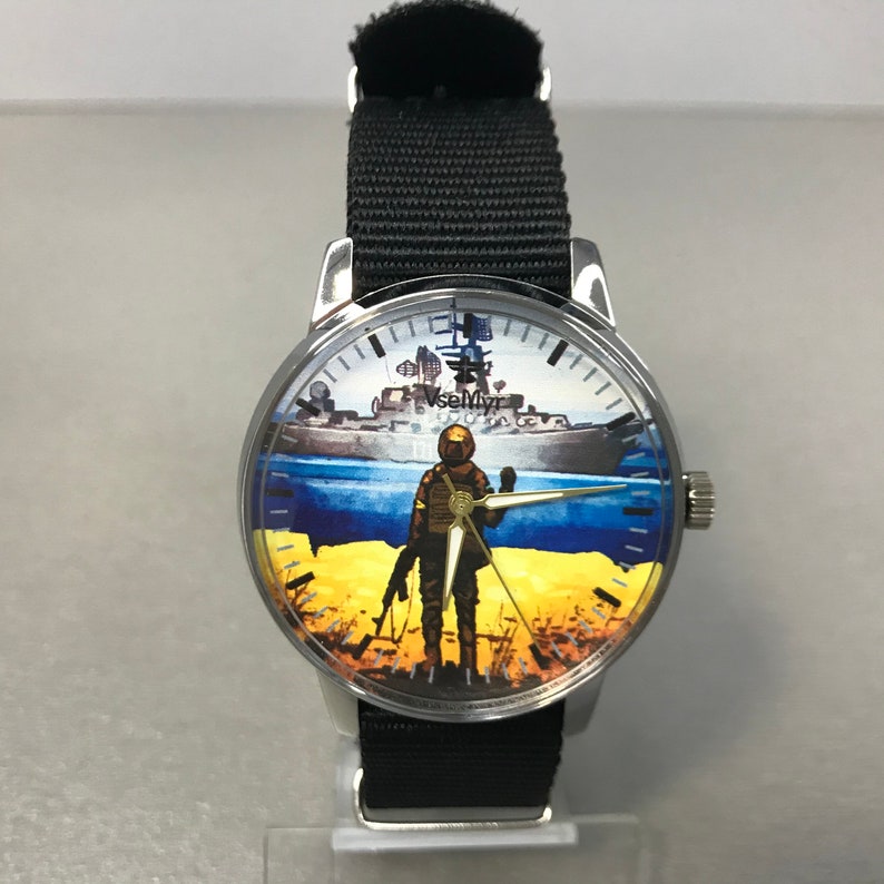 May include: A silver wristwatch with a black nylon band. The watch face features a soldier standing in front of a ship, with a yellow and blue background. The text "VseMyr" is visible on the watch face.