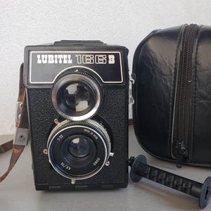May include: A black Lubitel 166B twin-lens reflex camera with a leather strap and a black case. The camera has two lenses and a film spool. The camera is made in the USSR.