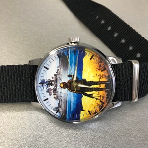 May include: A round wristwatch with a black nylon band. The watch face features a split design: a cityscape on the left and a soldier silhouette against a yellow and blue background on the right. The watch has silver-toned metal casing.