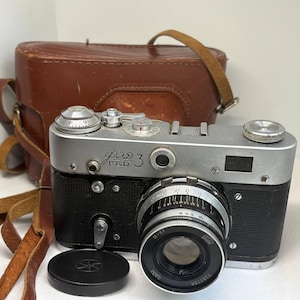 May include: A vintage FED 3 camera with a silver body and black accents, accompanied by a brown leather case and strap. The lens is black with silver details, and a black lens cap is in front. The camera has dials and settings visible.