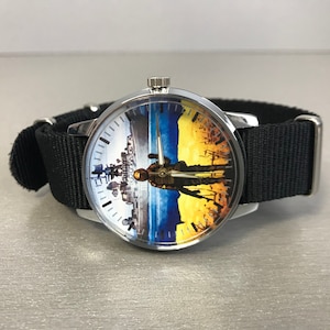 May include: A wristwatch with a black nylon band and a silver-colored case. The watch face features a split design with a cityscape on the left and a soldier silhouette against a yellow and blue background on the right. The watch hands are silver.