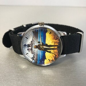 May include: A silver wristwatch with a black nylon band. The watch face features a split design with a cityscape on the left and a silhouette against a blue and yellow background. The text "VseMyz" is visible on the watch face.