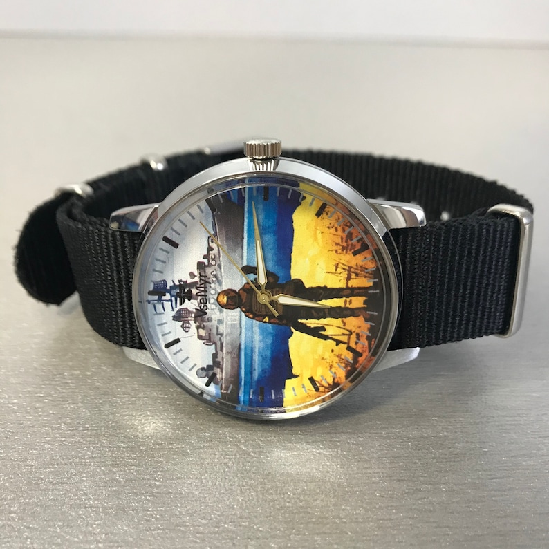 May include: A silver wristwatch with a black nylon band. The watch face features a split design with a cityscape on the left and a silhouette against a blue and yellow background. The text "VseMyz" is visible on the watch face.