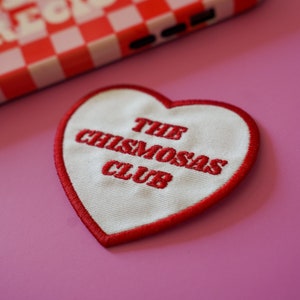 Puede incluir: Una funda de teléfono a cuadros rojos y blancos con las palabras "MORE PRECIOUS" en letras blancas. También se ve un parche en forma de corazón con el texto "THE CHISMOSAS CLUB" en bordado rojo sobre un fondo rosa.