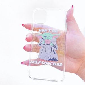 May include: Clear phone case featuring a green alien character holding a pink object, with the text "DON'T BE SELF CONCHAS" in white. The case is held against a white background.