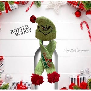 May include: A light green knitted bottle topper set featuring a beanie with a red pom-pom and a matching scarf. The beanie displays a black embroidered face, and the scarf features the red and green text Drink Up Grinches with a red wine glass.
