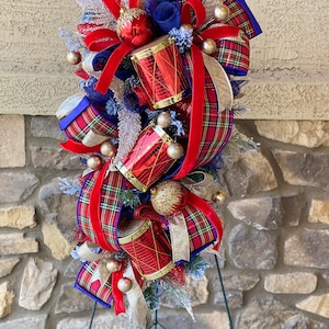 May include: A Christmas swag with red and blue plaid ribbon, red velvet ribbon, gold glitter ribbon, and red and gold drums. The swag is decorated with gold and red ornaments and is on a black metal stand.