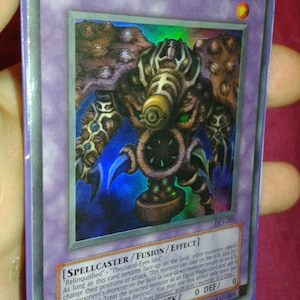 Könnte beinhalten: Eine Thousand-Eyes Restrict Sammelkarte aus dem Yu-Gi-Oh! Trading Card Game. Die Karte hat einen violetten Hintergrund mit einem dunkelblauen und grünen Monster mit mehreren Augen. Der Kartentext lautet "SPELLCASTER/FUSION/EFFECT" und "Relinquished" "Thousand-Eyes Idol".