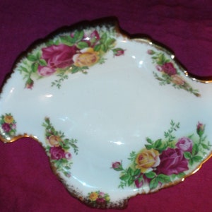May include: A white porcelain serving dish with a gold rim and a floral design featuring red, yellow, and green roses. The dish is shaped like a leaf and has a scalloped edge.