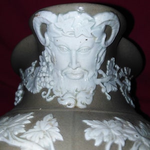 Könnte beinhalten: Eine Keramikkanne mit grüner Glasur und weißem Reliefdekor. Das Design zeigt ein Gesicht mit Hörnern und einem Kranz aus Blättern, Trauben und Ranken.