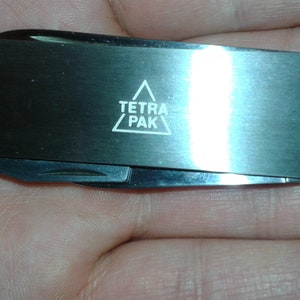 Puede incluir: Una multiherramienta plateada con un mango negro y un logotipo blanco que dice "TETRA PAK" en un triángulo.