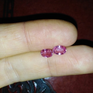 May include: Two pink oval-shaped gemstones, likely sapphires, are held between two fingers.