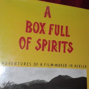 Op de afbeelding: Een gele boekomslag met de titel "A Box Full Of Spirits" in rode tekst. De ondertitel is "Adventures of a Film-Maker in Africa". De omslag toont ook een zwart-witfoto van een persoon die een televisie op zijn hoofd draagt, een geweer vasthoudt en door een veld loopt.