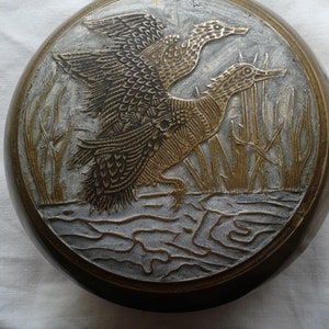 Puede incluir: Objeto metálico redondo, color bronce, con un relieve detallado de dos patos en vuelo sobre agua y juncos. Los patos están representados con intrincados detalles de plumas, sobre un fondo texturizado.