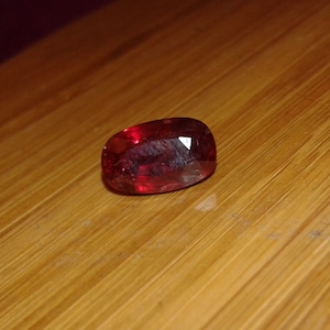 Puede incluir: Una piedra preciosa de color rojo intenso con forma ovalada facetada. La piedra preciosa está sobre una superficie de madera.