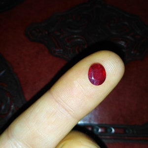 Puede incluir: Una gema roja intensa, facetada y de forma ovalada, posiblemente un rubí, descansa sobre un dedo. Las superficies pulidas de la gema reflejan la luz, mostrando su claridad y color intenso. El fondo es oscuro y texturizado.