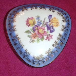 May include: A heart-shaped trinket dish with a blue and gold rim. The dish features a floral design with yellow, pink, and purple flowers.