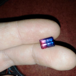 May include: A small, rectangular gemstone with a gradient of colors, including pink, purple, and blue. The gem is held between fingers, showcasing its polished facets and vibrant hues. The gem is likely a piece of jewelry.