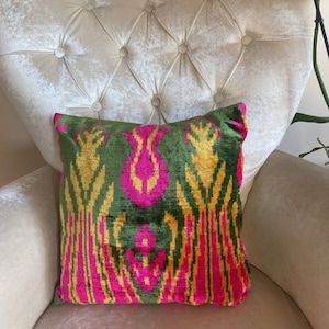 Può includere: Un cuscino da lancio verde, rosa e oro con un motivo e una texture in velluto. Il cuscino è seduto su una sedia bianca capitonné.