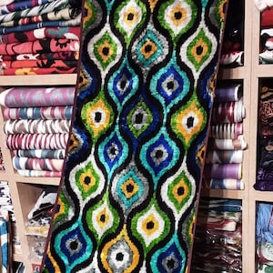 May include: A colorful fabric runner with a repeating pattern of teardrop shapes in shades of blue, green, yellow, and white. The fabric has a velvet-like texture and is displayed on a shelf with other fabrics.