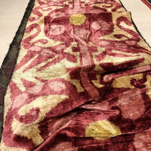 May include: A close-up of a burgundy and gold fabric with a repeating pattern. The fabric is folded and has a soft, velvety texture.