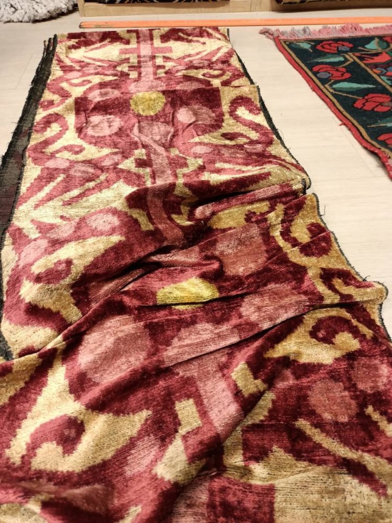 May include: A long, burgundy and gold patterned fabric with a velvet texture. The fabric is folded and draped on a light-colored surface.