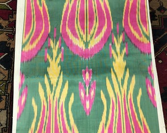 May include: A folded textile with a repeating pattern of stylized floral shapes in pink and yellow against a green background. The fabric has a silky sheen and is folded with white edges visible. The textile is likely used for clothing or home decor.