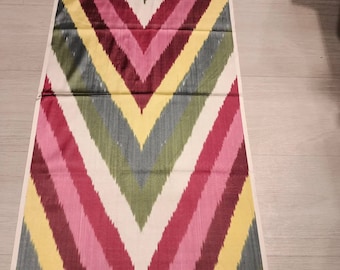 Può includere: Un tessuto colorato con un motivo a zig-zag. Il design presenta strisce alternate rosa, rosse, gialle, verdi, grigie e bianche. Il tessuto è disposto su una superficie chiara, mettendo in risalto i colori vivaci e il design geometrico.