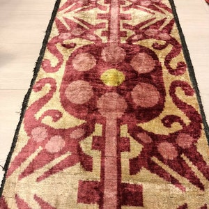 May include: A long, narrow rug with a repeating pattern of stylized flowers and geometric shapes. The rug is made of a soft, velvety fabric and has a rich, burgundy color with accents of gold and pink.