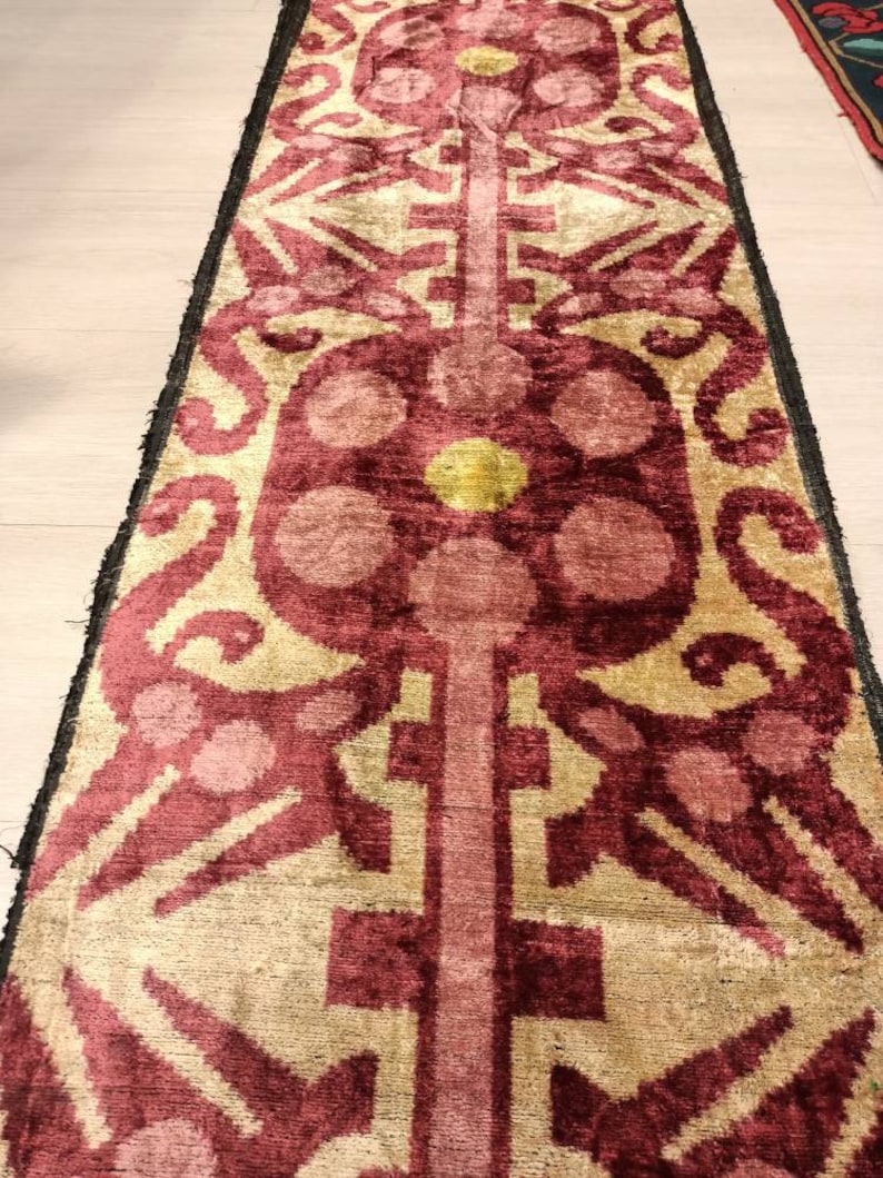 May include: A long, narrow rug with a repeating pattern of stylized flowers and geometric shapes. The rug is made of a soft, velvety fabric and has a rich, burgundy color with accents of gold and pink.