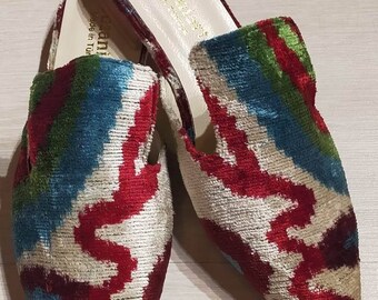 Könnte beinhalten: Ein Paar spitz zulaufende, offene Mules mit einem weißen, roten, grünen und blauen Ikat-Muster. Die Mules sind aus Samt und haben eine Lederinnensohle. Der Text "Made in Turkey" ist auf der Innensohle sichtbar.