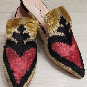 Puede incluir: Un par de zapatillas de terciopelo de punta puntiaguda con un diseño de corazón. Las zapatillas son negras, doradas y rojas con el texto "Serrani S. Made in Italy" en el interior de una zapatilla.