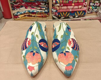 May include: A pair of white embroidered slippers with blue, green, pink, and red floral designs. The slippers have pointed toes and are made of a soft fabric.