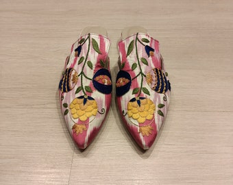 May include: A pair of pink and white embroidered slippers with floral designs. The slippers have a pointed toe and a flat heel.