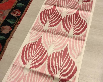 May include: A long, rectangular piece of fabric with a pink and red ikat pattern. The fabric has a white background with a repeating pattern of pink and red leaves.