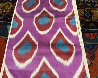 May include: A length of fabric with a repeating pattern of stylized teardrop shapes. The shapes are in shades of purple, red, and blue, outlined in white. The fabric is displayed on a patterned rug.