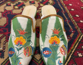 May include: A pair of embroidered slip-on shoes with a green and white striped pattern. The shoes feature floral embroidery in yellow, red, and blue. The trim is a dark brown color. The shoes are on a patterned red and blue rug.
