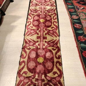 May include: A long, narrow rug with a repeating pattern of pink, yellow, and white. The rug has a traditional design with intricate details.