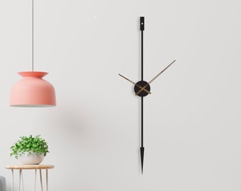 May include: A modern wall clock with long, thin hands and a small, round face. The clock is black and gold and is mounted on a white wall with a pink lamp hanging above it.