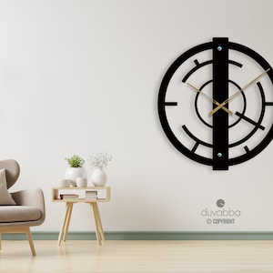 May include: Black metal wall clock with a unique geometric design. The clock has a large circular face with a vertical bar in the center. The clock hands are gold.