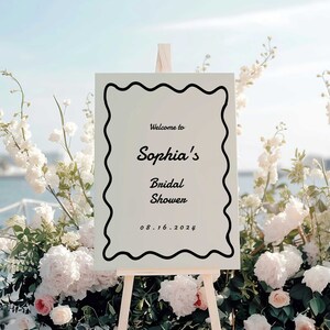 Könnte beinhalten: Ein weißes Schild mit schwarzem, wellenförmigem Rand, auf dem steht "Willkommen zum Bridal Shower von Sophia 08.16.2024". Das Schild steht auf einem Holzstaffelei vor weißen Blumen.