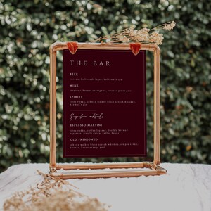 May include: A maroon sign with gold piping, listing the bar menu. The sign reads "THE BAR" and lists beer, wine, spirits, and signature cocktails. The signature cocktails are Espresso Martini and Old Fashioned.