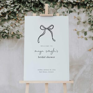 May include: A white sign with a black bow illustration and the text "WELCOME TO maya singh's bridal shower sunday, january 26, 2025 hart house, toronto".